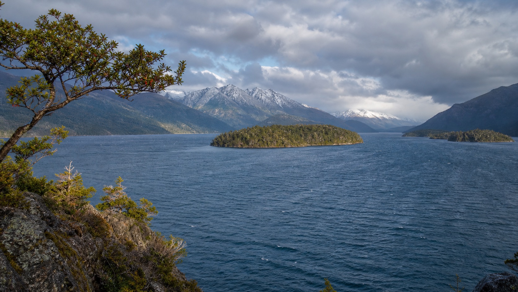 Lago Nahuel Huapi con islas boscosas y montañas nevadas bajo nubes de otoño